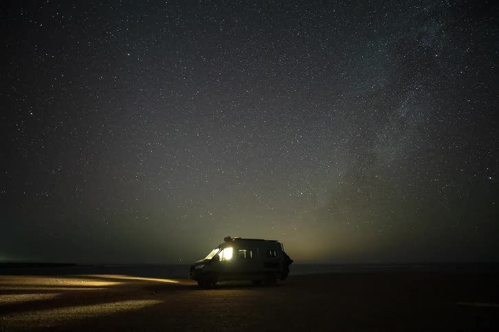 Stunning timelapse of the Milky Way and stars moving across the night sky above an illuminated camper van on a remote beach