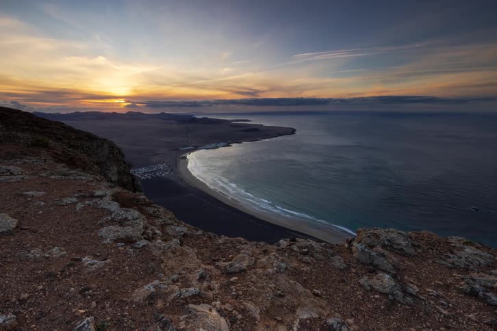 A stunning time-lapse from a cliffside viewpoint shows the sun setting over a vast bay and a dark sand beach