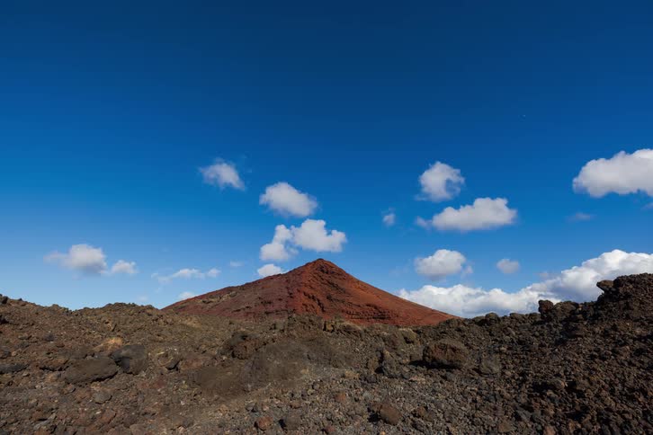 Timelapse of a red volcanic cone amidst a stark, rocky landscape with clouds moving across the vibrant blue sky