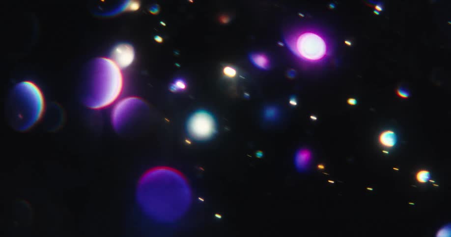 A magical display of vibrant, iridescent bokeh orbs and glittering particles float and drift through a dark space