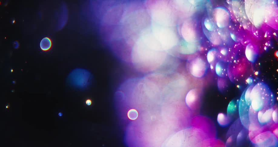 Colorful bokeh and shimmering particles drift slowly from a bright background into darkness, creating a magical and ethereal effect