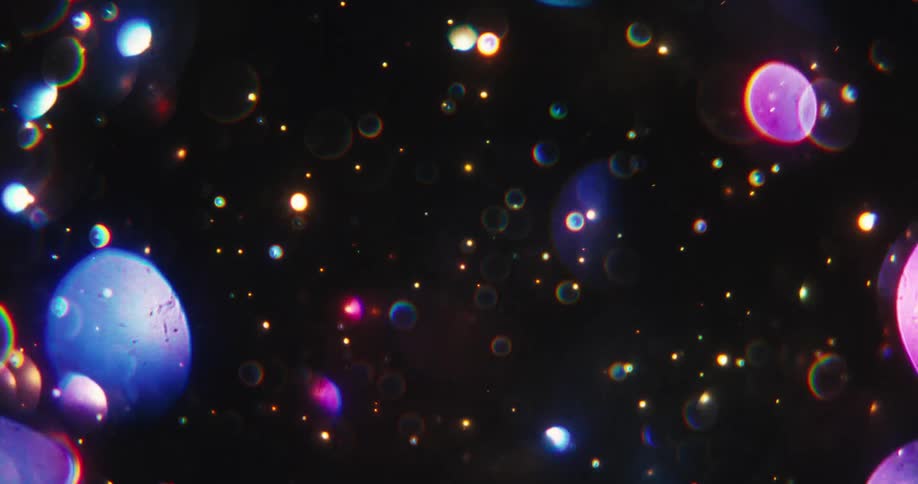 Colorful, dreamy bokeh orbs and sparkling particles float magically through the dark