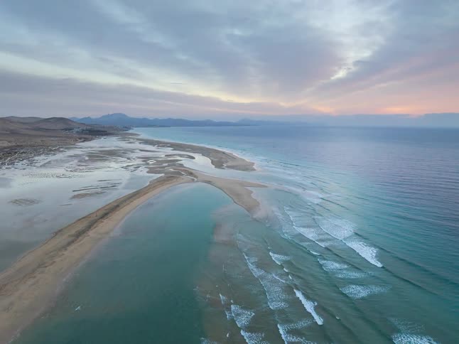 A stunning aerial view of a long sandbar stretching into the turquoise ocean under a colorful pastel sunset
