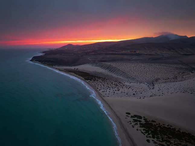 An epic aerial view captures a fiery red and orange sunset over a tranquil desert coastline meeting the sea