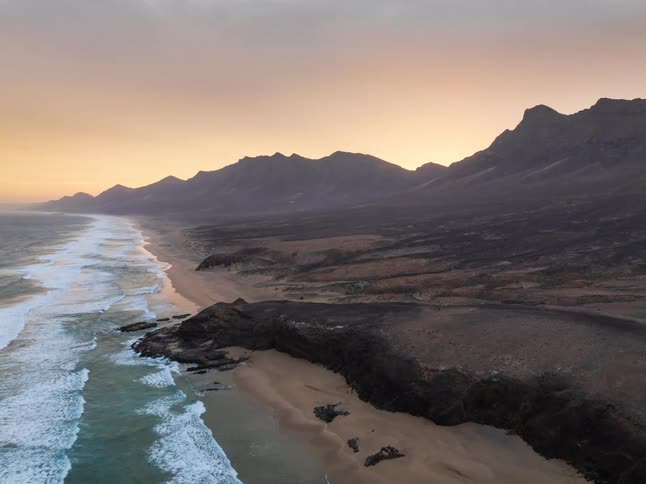 A stunning aerial shot slowly reveals a remote and rugged coastline with a sandy beach and mountain range during a beautiful golden sunset