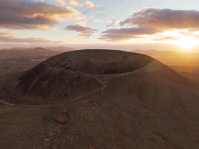 A dramatic aerial shot pulls back from a massive volcanic crater in a barren, rocky landscape as the sun sets on the horizon