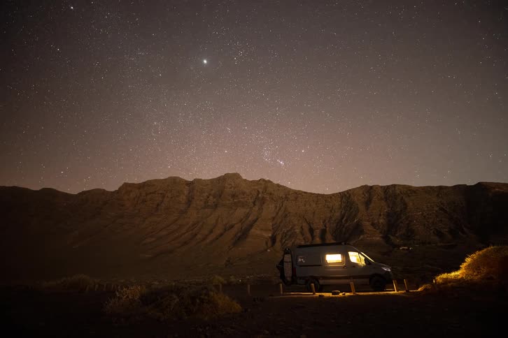 A cozy camper van provides a warm glow under a spectacular timelapse of the stars and Milky Way moving across a remote mountain desert