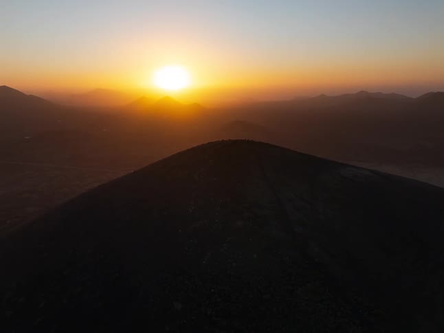 A slow aerial pullback reveals hikers silhouetted on a mountain peak against a beautiful, golden sunset