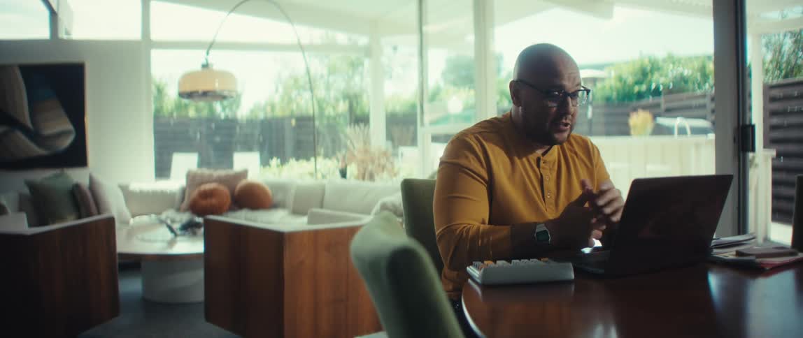 A professional man with glasses talks and gestures during a video meeting on his laptop in a modern, sunlit home