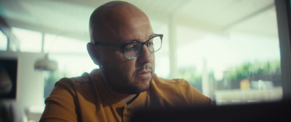 A serious bald man in glasses with a yellow shirt concentrates while working in a bright room
