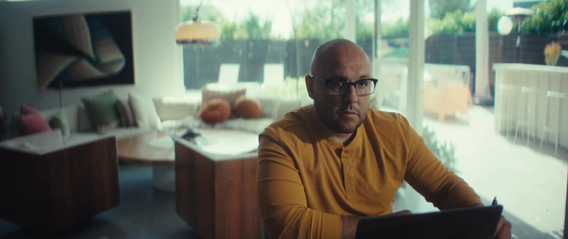 A man in a yellow shirt and glasses works on his laptop and takes notes in his modern, sunlit home office
