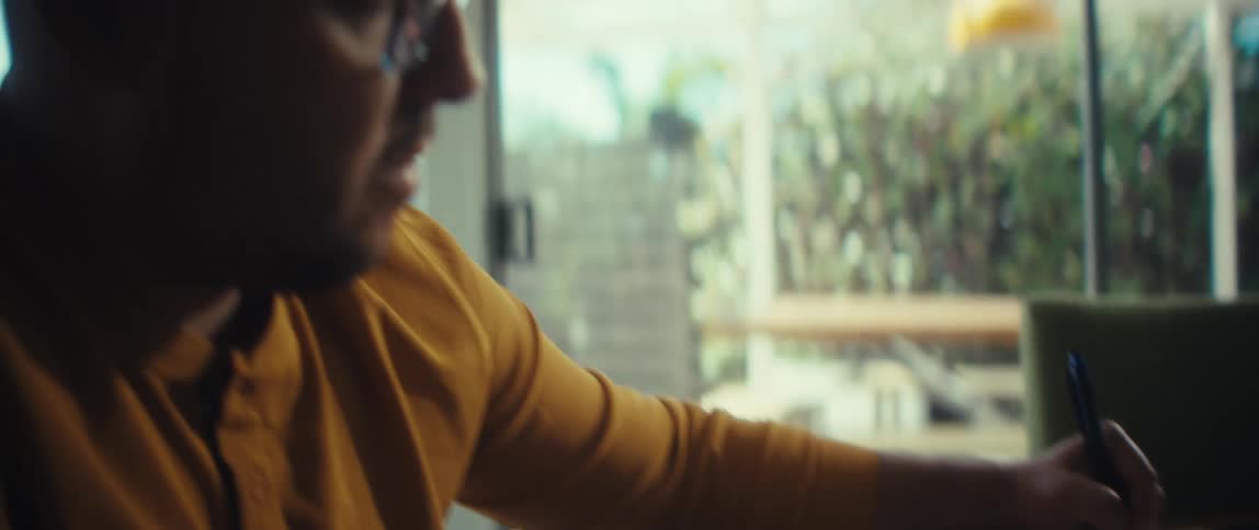 A focused man in glasses works from home, writing notes in a spiral notebook next to his laptop as the camera slowly zooms in on his face