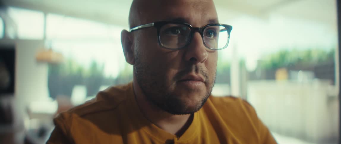 A focused man in glasses works from his home office, taking notes and looking at his laptop screen in a brightly lit room