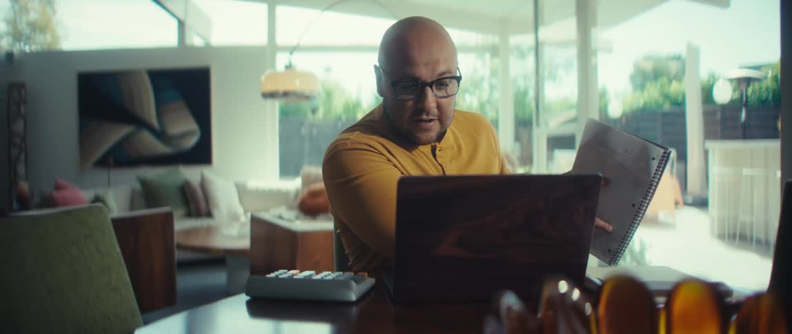 A bald man wearing glasses has a video conference meeting on his laptop while working from his modern home