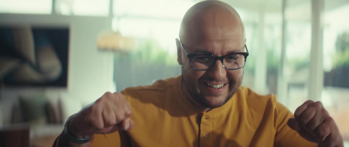 An ecstatic bald man wearing glasses celebrates a successful moment, smiling broadly and clenching his fists in a well-lit room