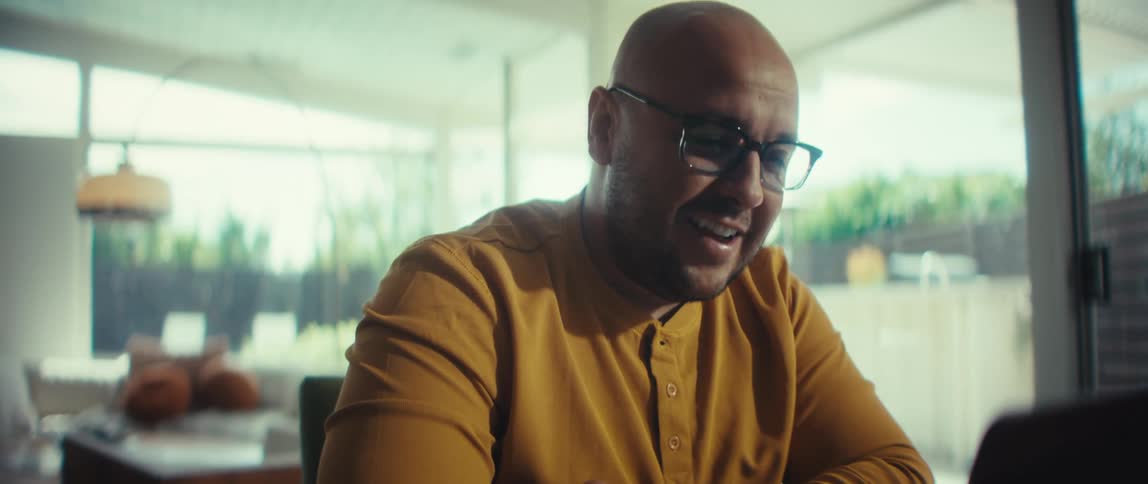 A cheerful bald man with glasses smiles and talks during a video conference call on his laptop in a modern home