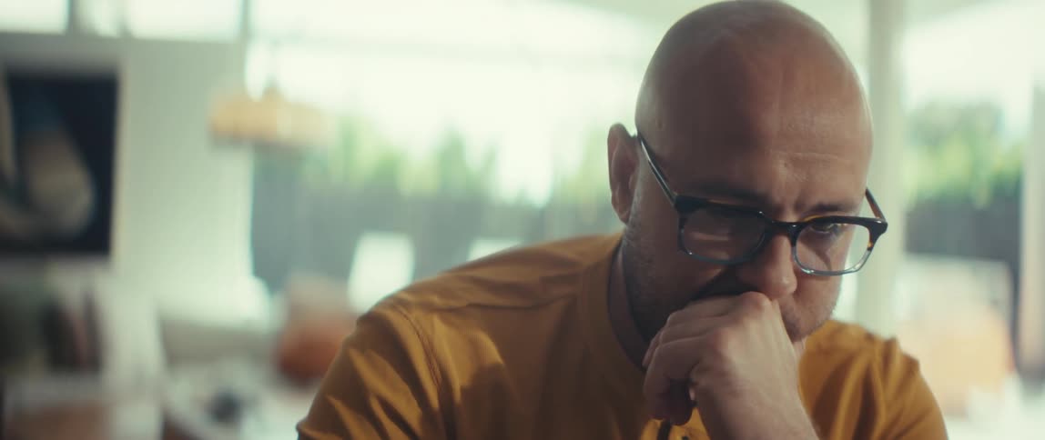 A pensive bald man with glasses in a yellow shirt sits in deep thought inside his modern home