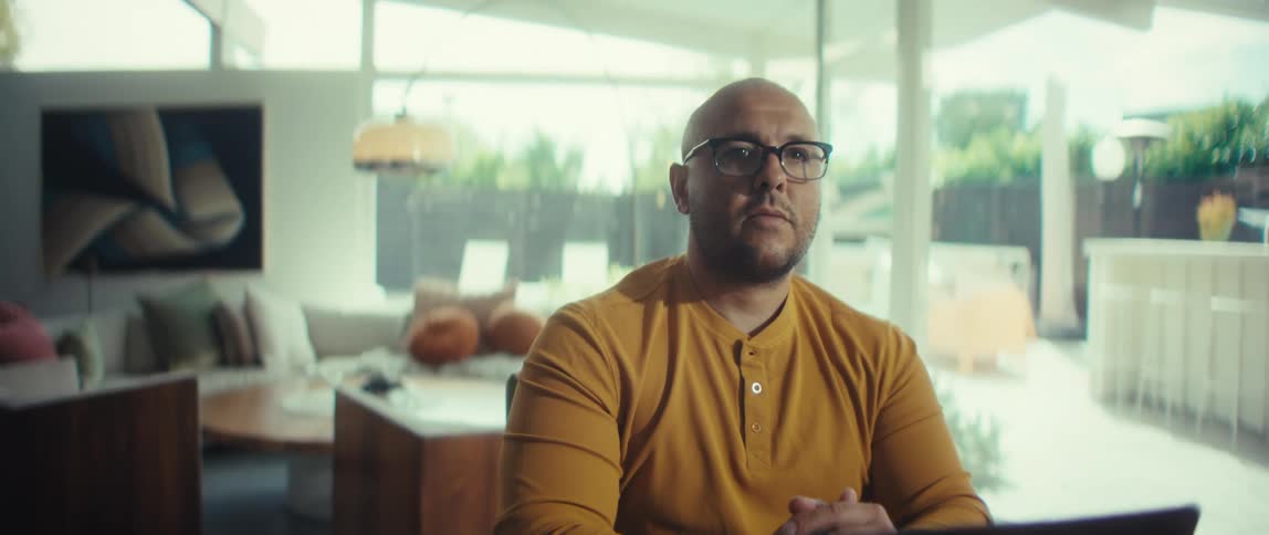 A serious man with glasses in a yellow shirt looks up from his work to stare at the camera in his modern, brightly lit home