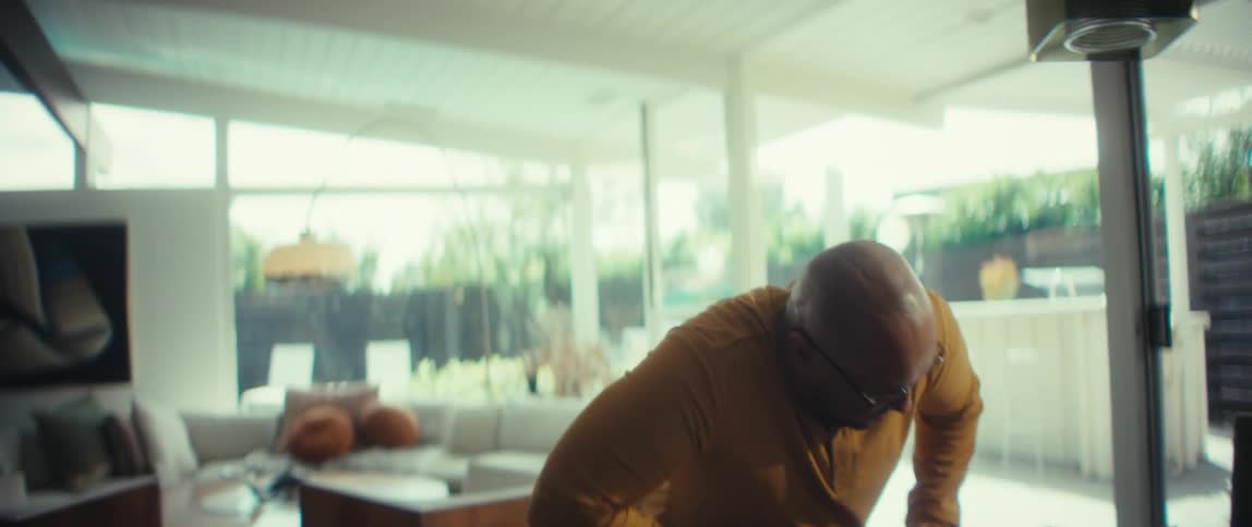 A bald man wearing glasses sits down at a dining table in a brightly lit, modern home, opens his laptop, and begins to work
