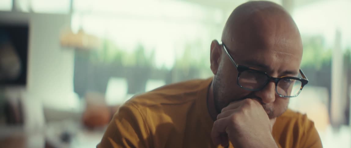 A close-up shot of a pensive bald man with glasses looking thoughtful and serious in a brightly lit room