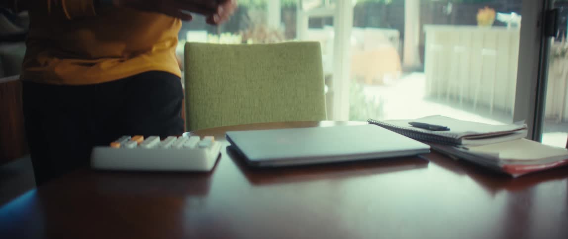 A man in a yellow shirt sits at a dining room table, sets down his phone, and opens his laptop to start working from home