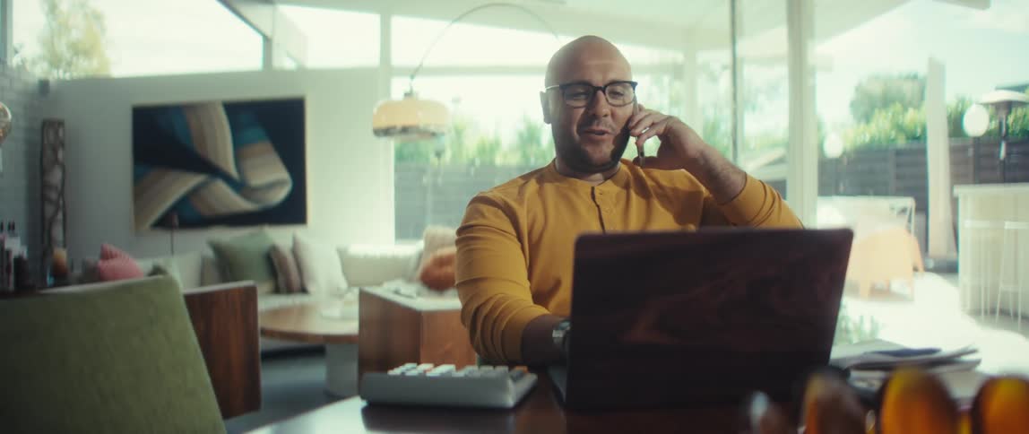 A professional man in a stylish, sunlit home office smiles as he answers a business call on his smartphone while working on his laptop