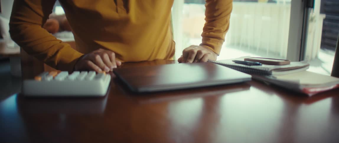 A man working from home in a sunlit room finishes his tasks, closes his laptop, and walks away to end his workday