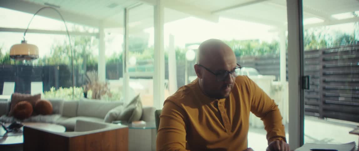 A bald man in a yellow shirt works from his stylish dining room, then closes his laptop and walks away, signaling the end of his workday