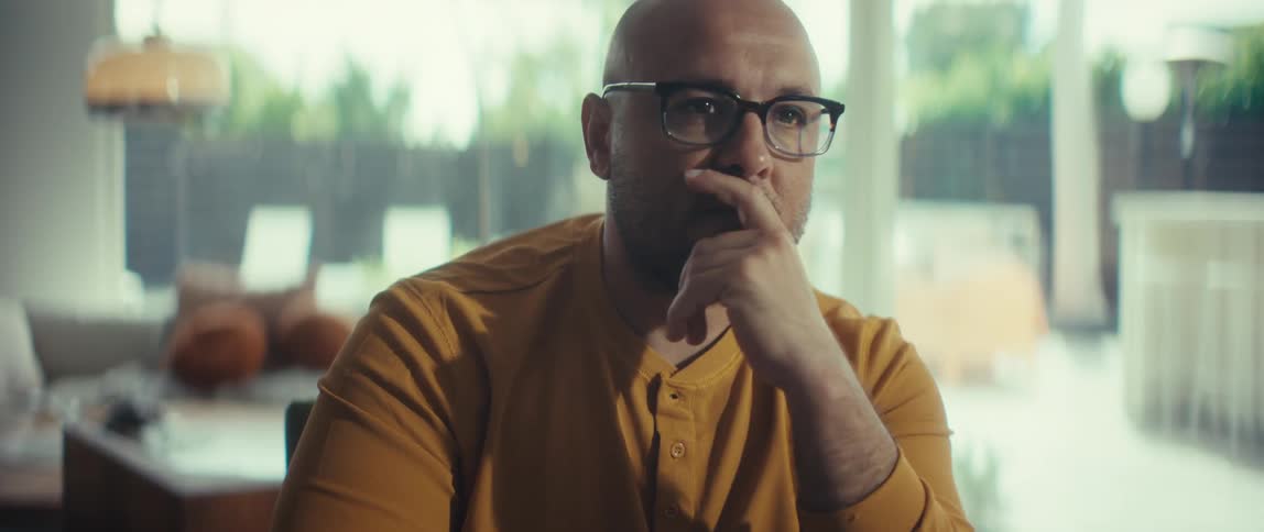 A focused bald man with glasses works intently on his laptop, rubbing his chin in deep thought in a brightly lit room
