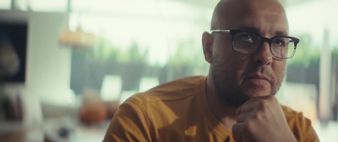 Close-up shot of a pensive bald man with glasses, deep in thought inside a modern, sunlit room