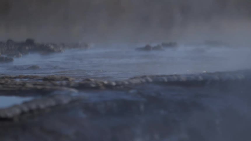 Steam gently billows across the surface of a dark, bubbling hot spring