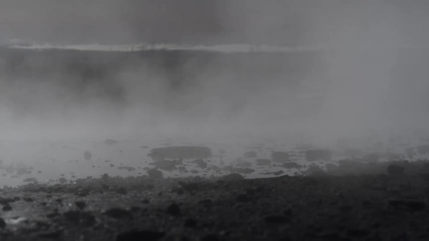 Silhouetted figures watch as thick steam rises from a geothermal hot spring in a desolate, rocky landscape