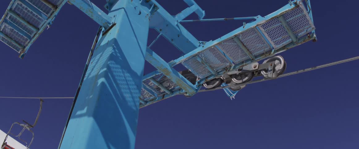 Low angle slow pan reveals the weathered details of a vintage blue ski lift tower against a clear sky