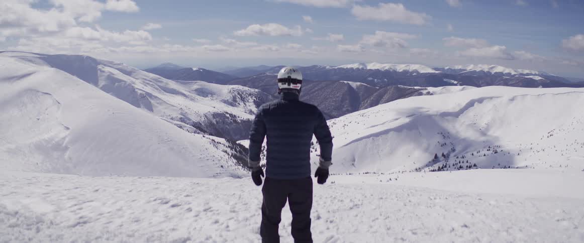 A skier stands on a mountaintop, looking out at a vast, snow-covered mountain range on a clear day