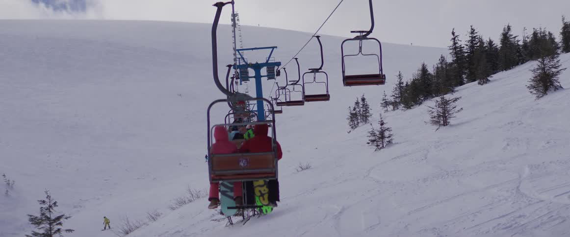 A stationary shot from behind shows a chairlift carrying people in winter gear up a snowy mountain slope at a ski resort