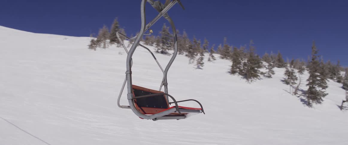 A point-of-view ride on a ski lift follows an empty chair up a snow-covered mountain on a sunny day