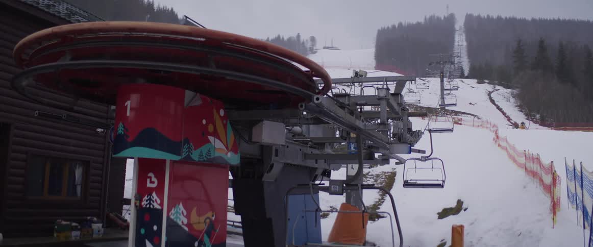 The mechanism of a ski lift turns as empty chairs ascend a snowy mountain on a cloudy day
