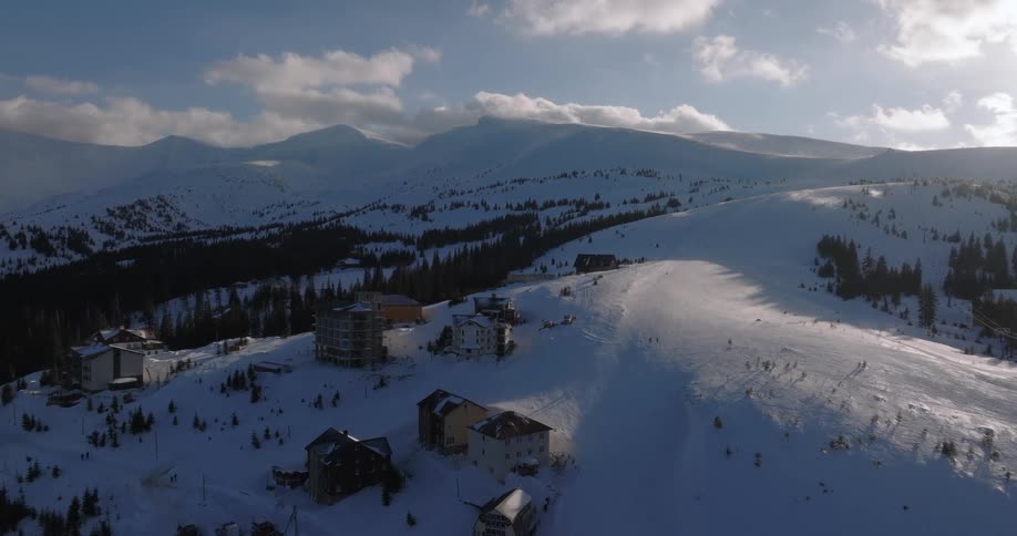 An aerial view captures a serene ski resort nestled in vast, snow-covered mountains under a partly cloudy sky