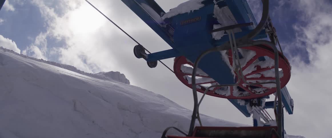 An empty ski lift chair moves in slow motion past a snow-covered pulley wheel on a sunny winter day