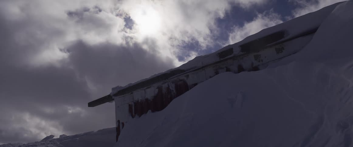 A red mountain hut is nearly buried in a deep snowdrift as wind blows snow under a dramatic, cloudy sky
