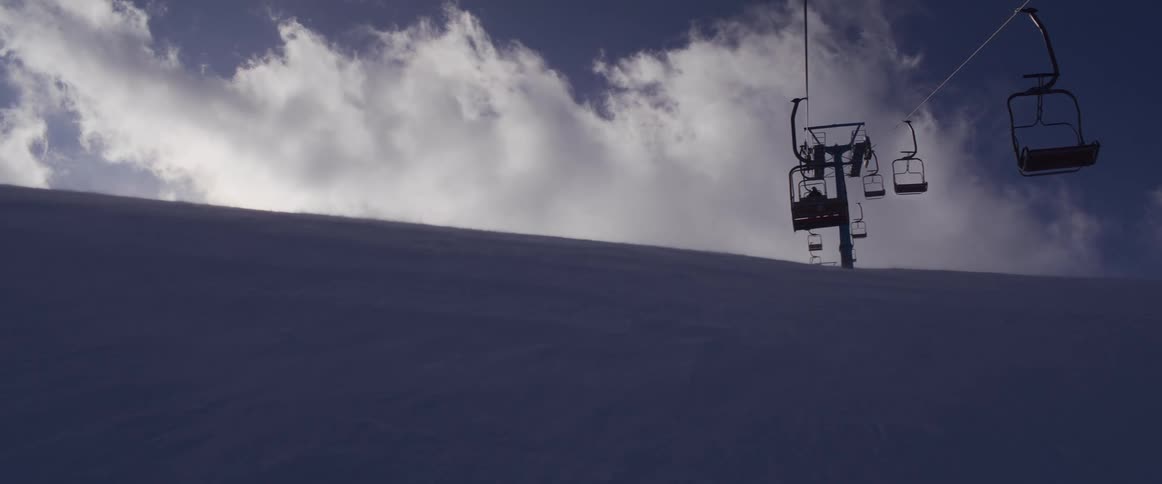 A ski lift carries passengers up a snowy mountain under a dramatic cloudy sky on a cold winter day