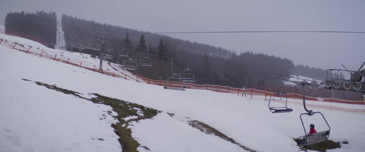 A ski lift carries empty chairs and a single skier up a snowy mountain slope on a quiet, overcast day