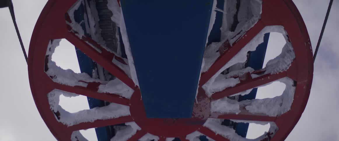 A close-up view of a large, colorful ski lift wheel encrusted with snow and ice, rotating slowly against an overcast sky