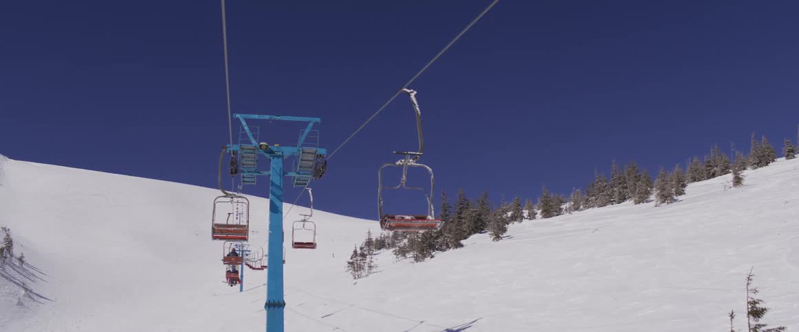 A ski lift in slow motion transports people up a snowy mountain under a clear blue sky on a sunny winter day