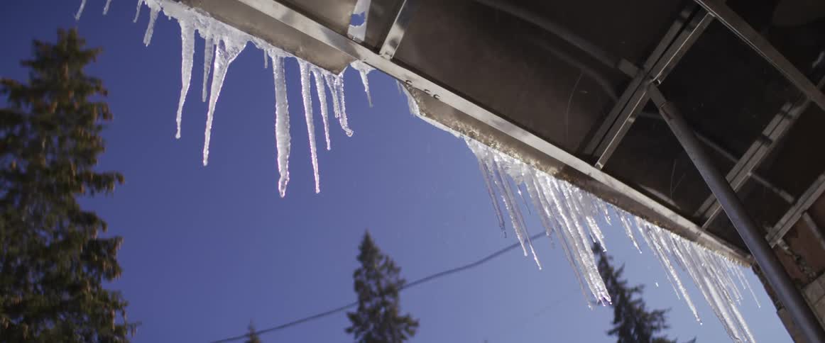 In slow motion, glistening icicles melt and drip from a roof's edge on a sunny winter day against a clear blue sky