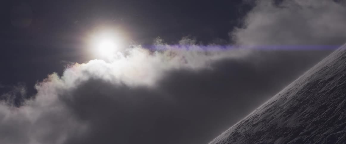 The sun shines through moody clouds as a fierce blizzard blows snow over a frozen mountain peak