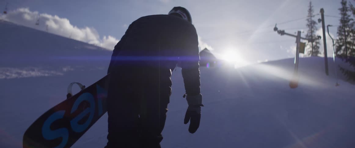 In slow motion, a snowboarder carries their board while walking up a snowy mountain slope towards the bright, setting sun