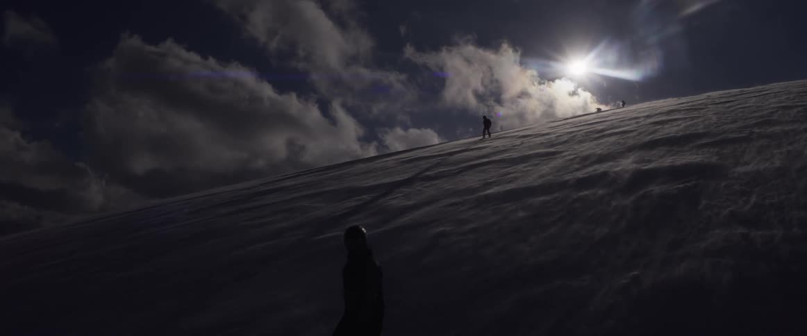 A dramatic slow-motion shot captures silhouetted snowboarders gliding down a vast, sun-drenched mountain of snow