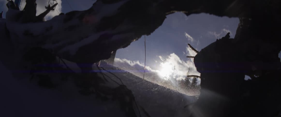 Wind whips sparkling snow across a sunny mountain landscape, viewed from a dark, snowy shelter in slow motion