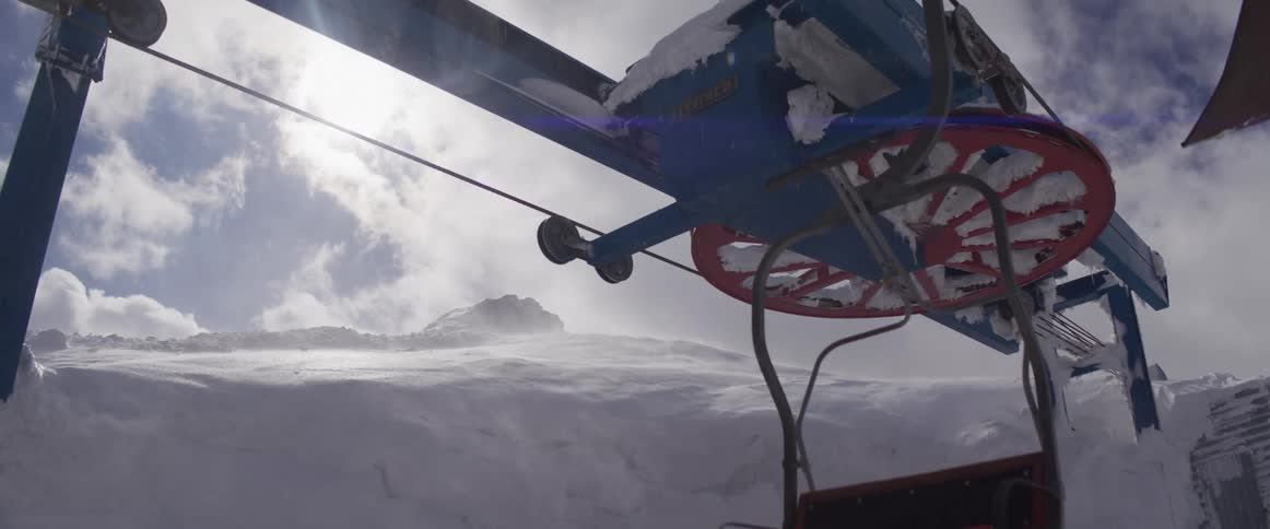 A ski lift bullwheel covered in snow and ice turns in slow motion as an empty chair passes by during a windy blizzard